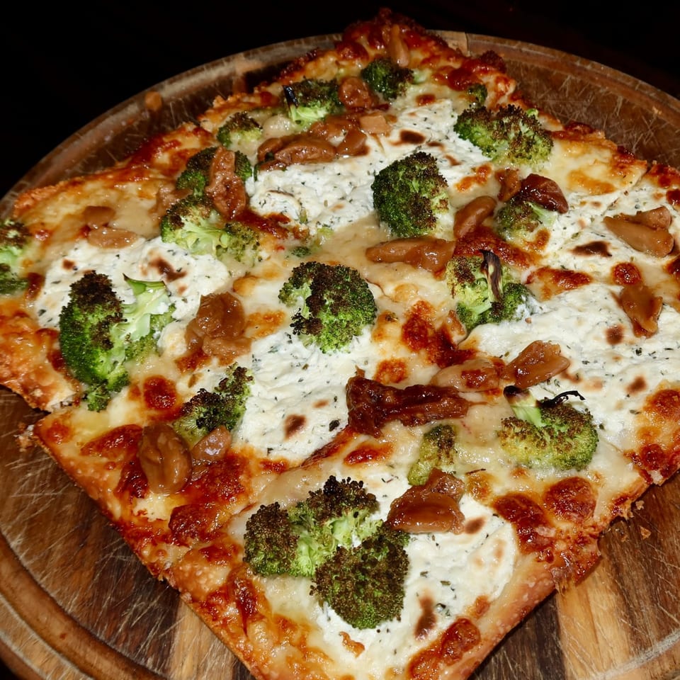 Square pizza with white cheese, broccoli florets, and mushrooms on a wooden board.