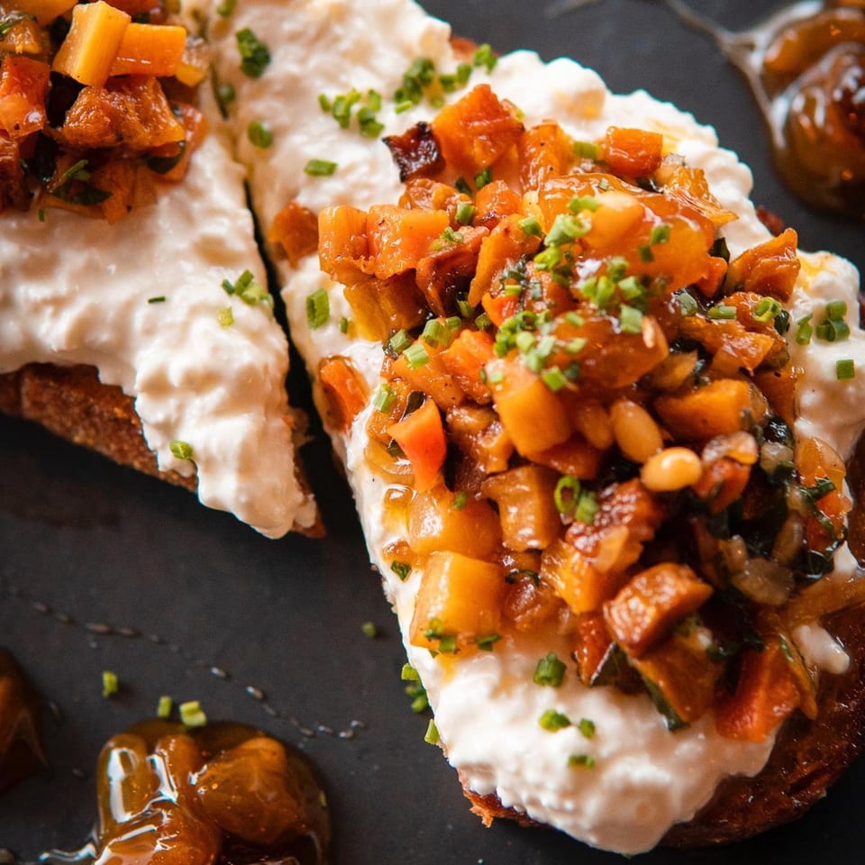 Two pieces of toast topped with creamy white cheese, diced orange vegetables, and green chives.