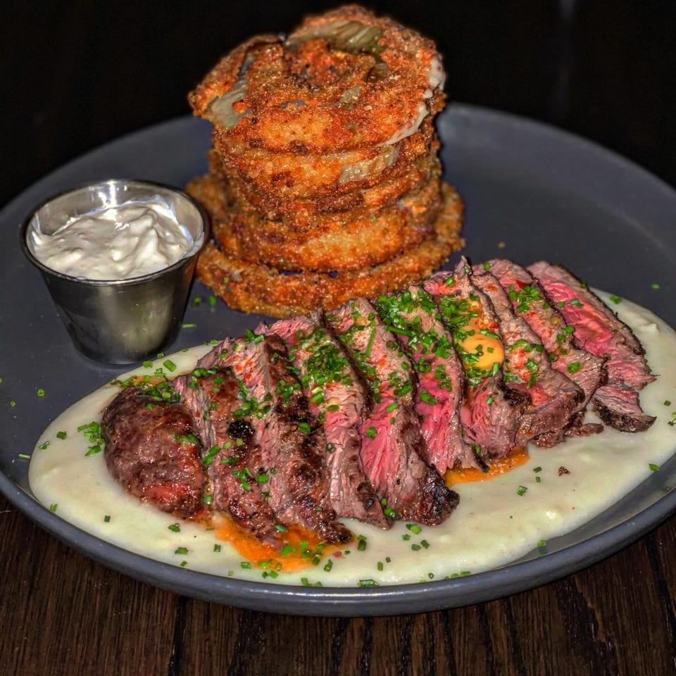 Sliced steak on mashed potatoes with chives, a side of fried onion rings, and dipping sauce.