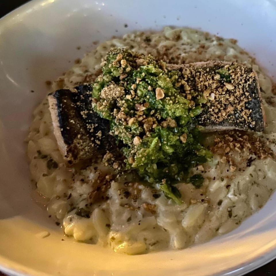 Seared salmon with green pesto and crushed nuts on a bed of creamy risotto in a white bowl.